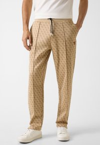 Beige joggers met een patroon en een textuurontwerp met rode accenten, voorzien van een tailleband met trekkoord en zijzakken, gecombineerd met witte sneakers.