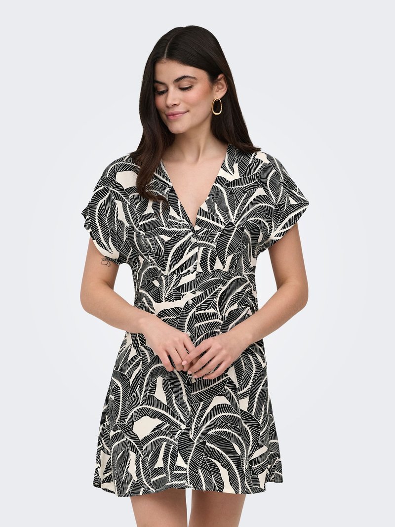 Femme en robe à manches courtes, motif feuilles noir et blanc, col en V et boucles d'oreilles créoles dorées, debout devant un fond clair uni.