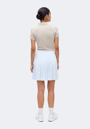 Kvinde med mørkt hår i en knold iført en beige kortærmet polo, hvid plisseret nederdel, hvide sokker og hvide sneakers, stående med ryggen mod os.