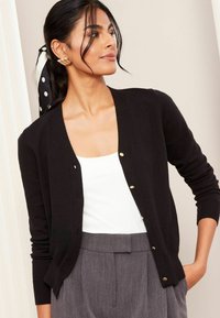 Cardigan negru cu nasturi aurii, cu manșete și tiv ribbated, purtat peste un top alb și pantaloni gri cu talie înaltă, dintr-un material texturat.