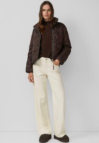 Braune gesteppte Jacke mit hohem Kragen, cremefarbene, weit geschnittene Hosen und dunkle Schuhe. Mit Reißverschluss und Vordertaschen.