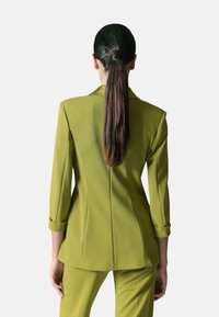 Blazer verde oliva con spalle strutturate, vestibilità sartoriale e maniche arrotolate; realizzato in tessuto liscio, con cuciture verticali per dare forma.