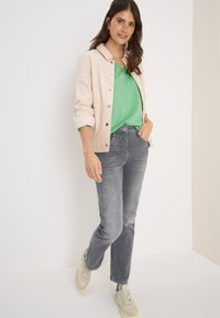 Vaalean beige corduroy-takki, vihreä V-kaula-aukko neule, harmaat farkut ja beige lenkkarit. Luonnollinen rakenne, yksinkertainen muotoilu, ei kuvioita tai koristeita.