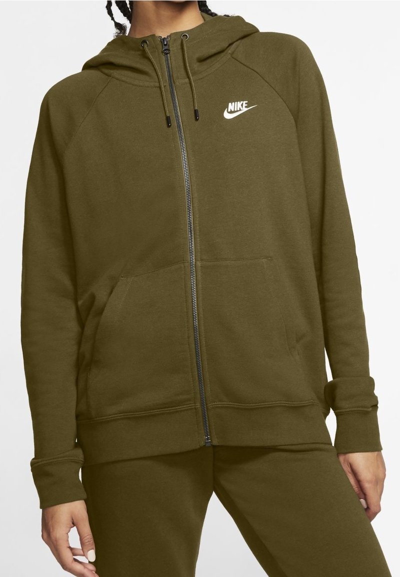 Femme portant un sweat à capuche zippé Nike vert olive avec poches avant et pantalon assorti, debout devant un fond blanc.