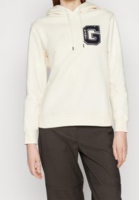 Sudadera con capucha crema con bolsillo frontal, que presenta un logo bordado en azul oscuro. Tejido suave con cordones y puños acanalados. Combinada con pantalones oscuros.