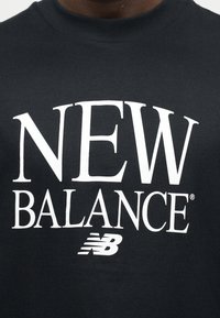 Κοντινό πλάνο ενός σκούρου πουκάμισου με μεγάλο λευκό κείμενο "NEW BALANCE" και το λογότυπο "NB" της μάρκας στο κέντρο του στήθους.