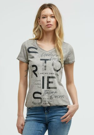 Woman with long blonde hair wearing a gray graphic t-shirt and blue jeans, standing against a plain light background.