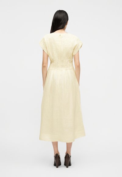 Γυναίκα με μακριά σκούρα μαλλιά, φορώντας κρεμ υφασμάτινο μίντι φόρεμα και σκούρα καφέ ψηλοτάκουνα παπούτσια, στέκεται με την πλάτη προς το μέρος μας σε απλό φόντο.