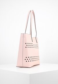 Lichtroze totebag met een gestructureerd ontwerp, reliëfstructuur, lange handgrepen en decoratieve perforaties aan de voorkant.