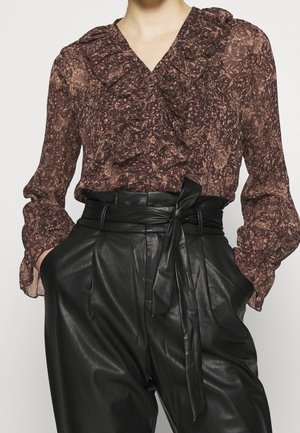 Brun mønstret bluse med flæser kombineret med højtaljede sorte læderpaperbagbukser bundet i taljen med et bælte.