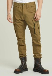 Olijfgroene cargobroek met meerdere zakken, ritssluitingen en taps toelopende pijpen. Gemaakt van stevig materiaal, gecombineerd met zwarte leren laarzen.