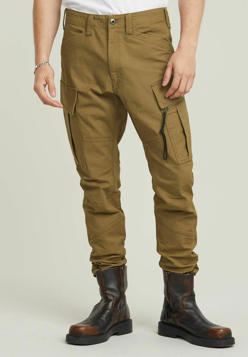 Olijfgroene cargobroek met meerdere zakken, ritssluitingen en taps toelopende pijpen. Gemaakt van stevig materiaal, gecombineerd met zwarte leren laarzen.