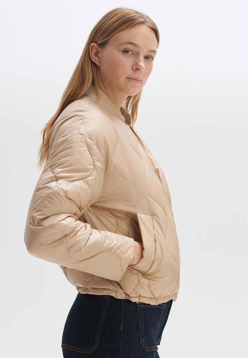 Beige gesteppte Bomberjacke mit geripptem Kragen, seitlichen Taschen und glatter, glänzender Textur. Modell steht im Profil vor einem schlichten Hintergrund.