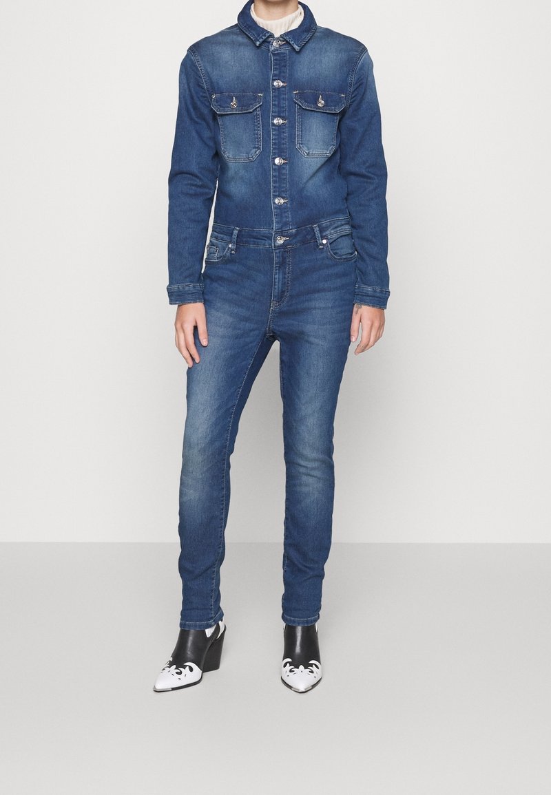 Personne portant une veste en jean bleue cintrée et un jean assorti, associée à des bottines à talons pointues noires et blanches, debout devant un fond uni.
