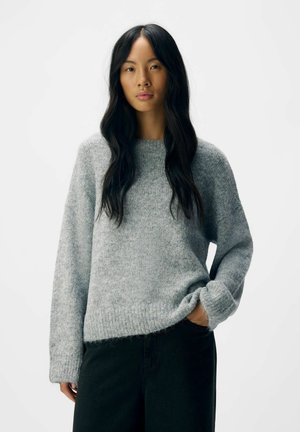 Gray knit sweater with a round neckline and wide sleeves, featuring a soft, textured finish. Paired with dark pants.