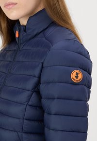 Marineblauwe pufferjas met een hoge kraag, een gequiltde textuur en een oranje accentknop met een logo op de linkermouw.