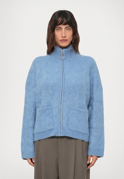 Woman wearing a light blue cable-knit zip-up sweater with front pockets and a ring zipper pull, paired with brown wide-leg pants.