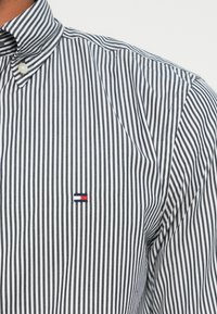 Stripet skjorte i navy og hvit bomull, med knappekrage. Brodert logo i rødt, hvitt og navy nær brystet.