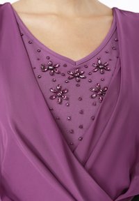 Lilla plagg med v-hals design dekorert med blomstertrykk laget av perler og edelstener, som viser en glatt stofftekstur.