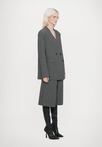 Blazer grigio su misura con doppio bottone, abbinato a pantaloni corti coordinati e stivali neri al ginocchio. Tessuto liscio e vestibilità strutturata.