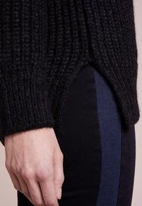 Schwarzer Strickpullover mit gerippter Textur, ausgestattet mit einem Seitenschlitz, kombiniert mit dunklen Hosen, die an der Seite einen blauen Streifen haben.