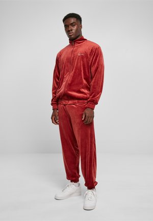 Survêtement en velours rouge rouille avec un col haut, une fermeture éclair et des poignets élastiques ; associé à des baskets blanches. Texture lisse, design minimaliste.