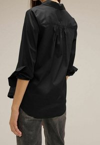 Camicia nera di cotone con maniche lunghe, polsini volant e dettagli di pieghe nella parte posteriore. Tessuto liscio e design del colletto classico.