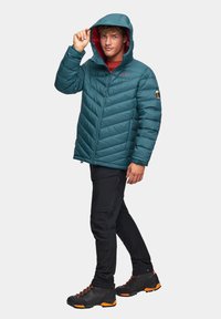 Turkusowa kurtka puchowa z kapturem, z charakterystycznym pikowaniem w zygzak. Nosi ją osoba w czerwonej bluzce, zestawionej z czarnymi spodniami i solidnymi butami trekkingowymi z pomarańczowymi akcentami.