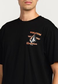Sort bomulds-t-shirt med grafisk design, der viser "Volcom" tekst i orange og hvid, et diamantlogo og ordet "Workwear". Casual pasform.