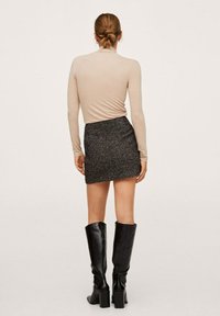 Beige lange mouw top gecombineerd met een gestructureerde zwarte mini rok en hoge zwarte hakken, gezien vanaf de achterkant.