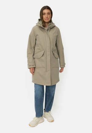 Kvinde iført en beige hættejakke til vinteren, blå jeans og hvide sneakers, der står med neutralt ansigtsudtryk mod en ensfarvet baggrund.