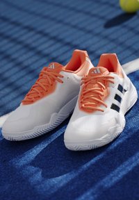 Λευκά και πορτοκαλί παπούτσια τένις Adidas τοποθετημένα σε μπλε επιφάνεια γηπέδου με σκιά τενιστικής μπάλας στο φόντο.
