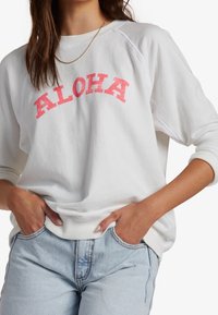 Kvinna iklädd en vit sweatshirt med "ALOHA" i rosa bokstäver, tillsammans med ljusblå jeans, händerna nedstoppade i framfickorna.