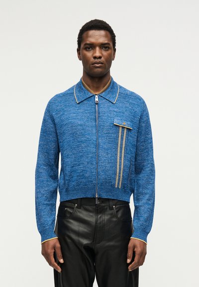 Άνδρας που φοράει μπλε πουλόβερ με φερμουάρ, κίτρινη επένδυση και κάθετη τσέπη με σχέδιο, συνδυασμένο με μαύρο δερμάτινο παντελόνι, στέκεται μπροστά από απλό φόντο.