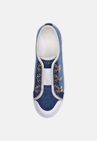 Denim slip-on tennarit, joissa on valkoinen kuminen kärki, kultaiset ketjureiät ja joustava nauha, jonka sisäpohjassa on "GUESS" -merkintä.