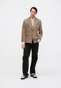 Beige korduroy blazer s dvojitým zapínáním, šedá košilová košile, tmavé kalhoty a černobílé boty. Ozdobné knoflíky a kapsy.