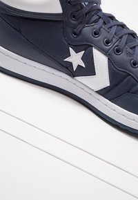 Vysoké tenisky v námořnické plátno s bílými koženými akcenty, se značkou hvězdy a kontrastní podrážkou. Šněrovány námořnickými tkaničkami.