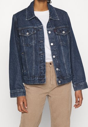 Personne portant une veste en jean bleu foncé sur un t-shirt blanc et un pantalon en velours côtelé beige, debout avec les bras détendus le long du corps.