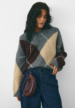 Prevelik pulover z argyle vzorcem v sivi, rjavi in bež barvi ter belimi detajli. Združen z temno modrimi kavbojkami in burgundskim nahrbtnikom.