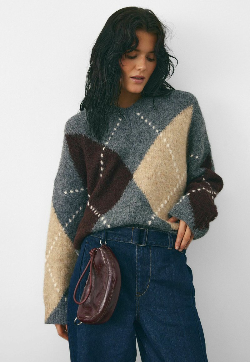 Per didelėme megztinyje su pilka, ruda ir smėlio spalvos argyle raštu, akcentuojama baltomis detalėmis. Suderinta su tamsiai mėlynomis džinsomis ir bordo spalvos rankine.