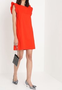 Czerwona sukienka bez rękawów z falbaniastymi ramionami, połączona z torebką w czarno-białym wzorze oraz srebrnymi czółenkami z ostrym noskiem.