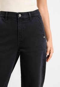 Mujer con jeans negros de cintura alta, con botón en la parte delantera y lazos para cinturón, de pie frente a un fondo blanco liso.