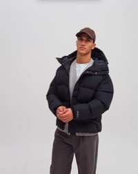 Junger Mann trägt eine schwarze Steppjacke, graue Hose, weißes Hemd und braune Mütze und steht vor einem schlichten grauen Hintergrund.