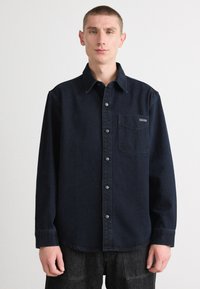 Camisa de denim azul escuro com mangas longas, colarinho pontiagudo, fecho com botões, bolso frontal com patch de logo e ajuste descontraído. Tecido texturizado.