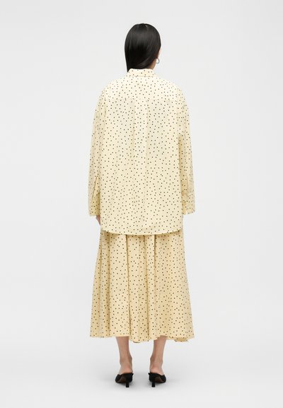 Γυναίκα με μακριά μαύρα μαλλιά, φοράει μια κρεμ μακρυμάνικη μπλούζα και πλισέ φούστα με μαύρα πουά, μαύρα mules με τακούνι, στέκεται με την πλάτη προς τα μέσα.