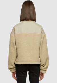 Žena sa ravnom smeđom kosom nosi bež flis jaknu s glatkom gornjom stražnjom panelom, uparenu s crnim hlačama, okrenuta leđima.