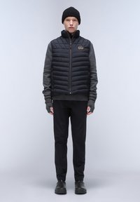 Schwarze Steppweste mit gestepptem Muster, graues Langarmshirt, schwarze Hose und schwarze Mütze. Mit Reißverschluss und Fäustlingen ohne Finger.