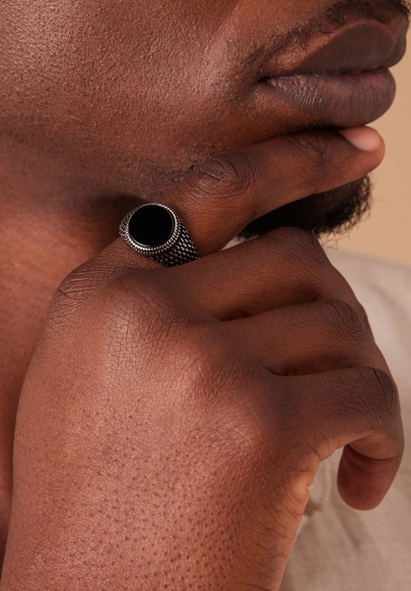 Schwarzer Steinring mit strukturiertem silbernen Netzband, getragen an einer Hand, die nahe dem Gesicht ruht, und das detaillierte Handwerk und Design hervorhebt.