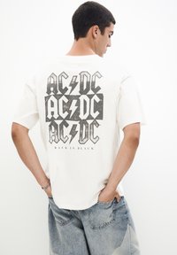 Krémové tričko s krátkým rukávem, na zadní straně s velkým černým grafickým designem s nápisem "AC/DC" a motivy blesků. Uvolněný střih.
