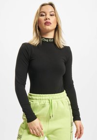 Langermet svart bodysuit med høy krage som har "Thug Life" i lys grønn. Kombinert med lys grønne joggebukser med side mønster.